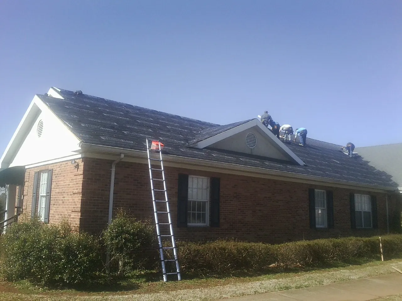 Crew tearing shingles off a brick ranch roof with a ladder set — Metal Roof Repair work by our crew in East Smithfield