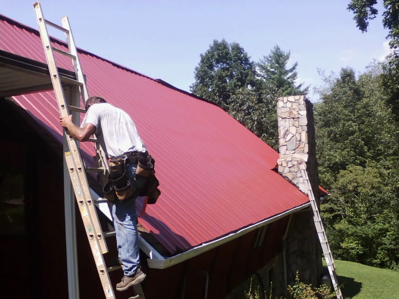 Roofer on a ladder beside a red metal roof and stone chimney — Soffit & Fascia Repair by Roofing Brothers in East Smithfield
